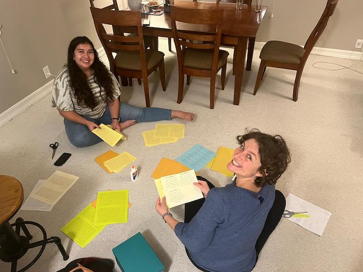 Photo of two smiling persons, working on the floor, surrounded by colored papers, envelopes, scissors and glue.